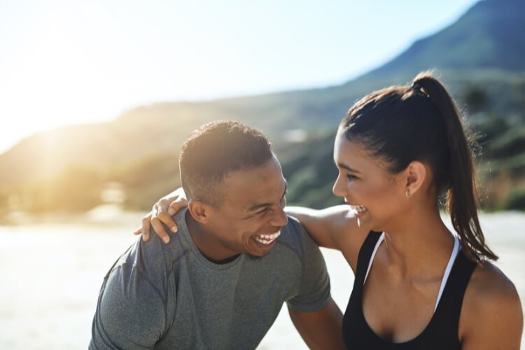 Casal sorrindo com vista de mar e montanhas ao fundo o sol reflete atrás deles
