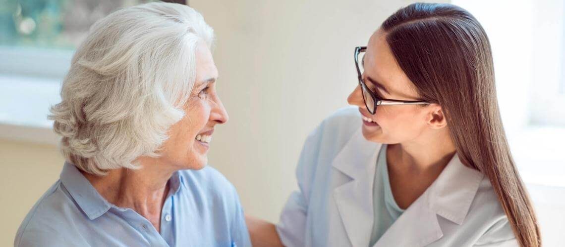 Senhora de cabelo branco sorrindo ao lado de médica que também está sorrindo para ela